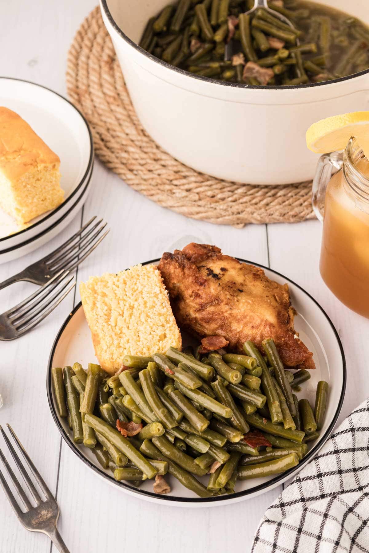 A plate of southern green beans with bacon, fried chicken, cornbread, and sit on a white wooden table, sweet tea and the pot of beans in the background.