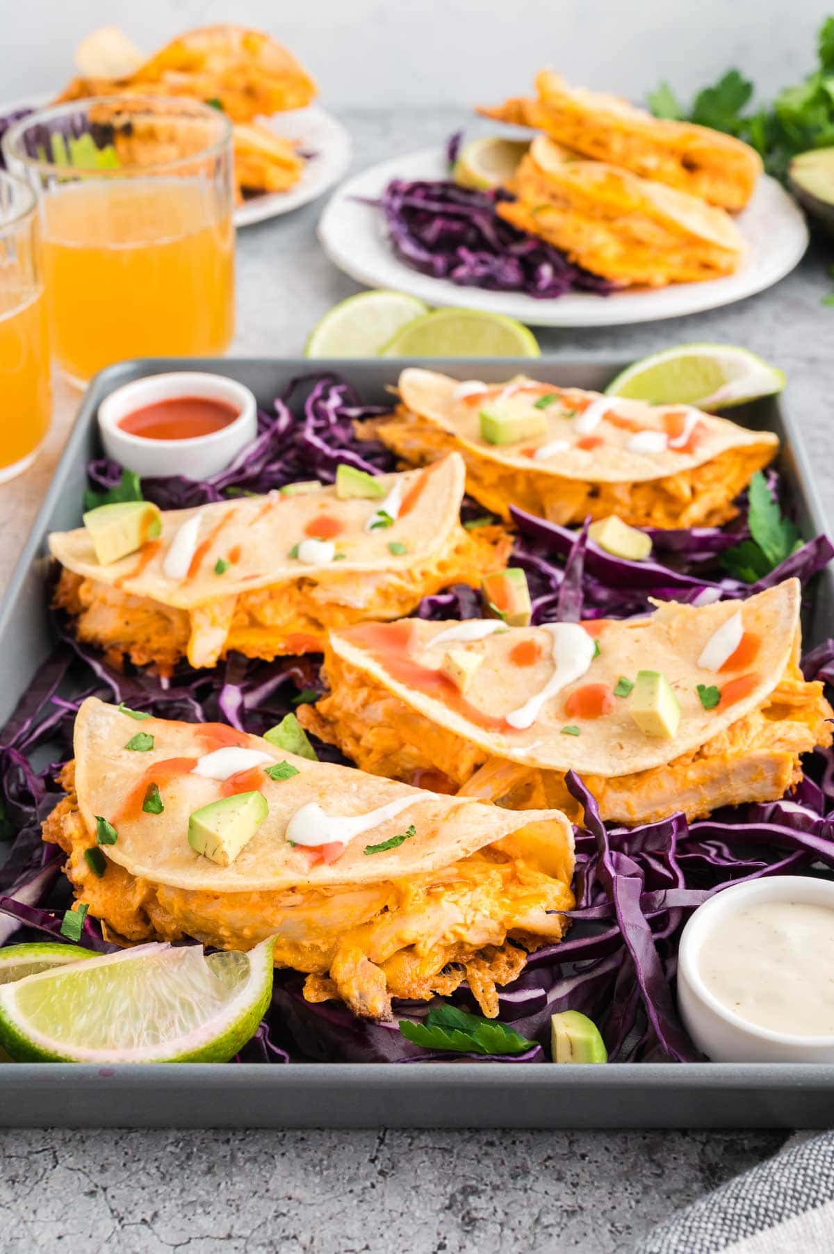 Four buffalo chicken tacos served on a small sheet pan with red cabbage, lime, and sauces.