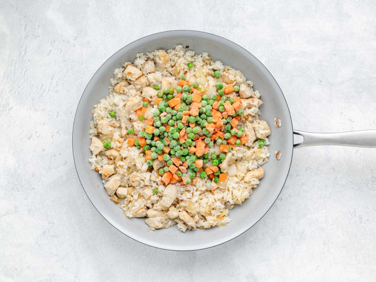 A skillet with diced chicken, rice, onion, peas, and carrots.