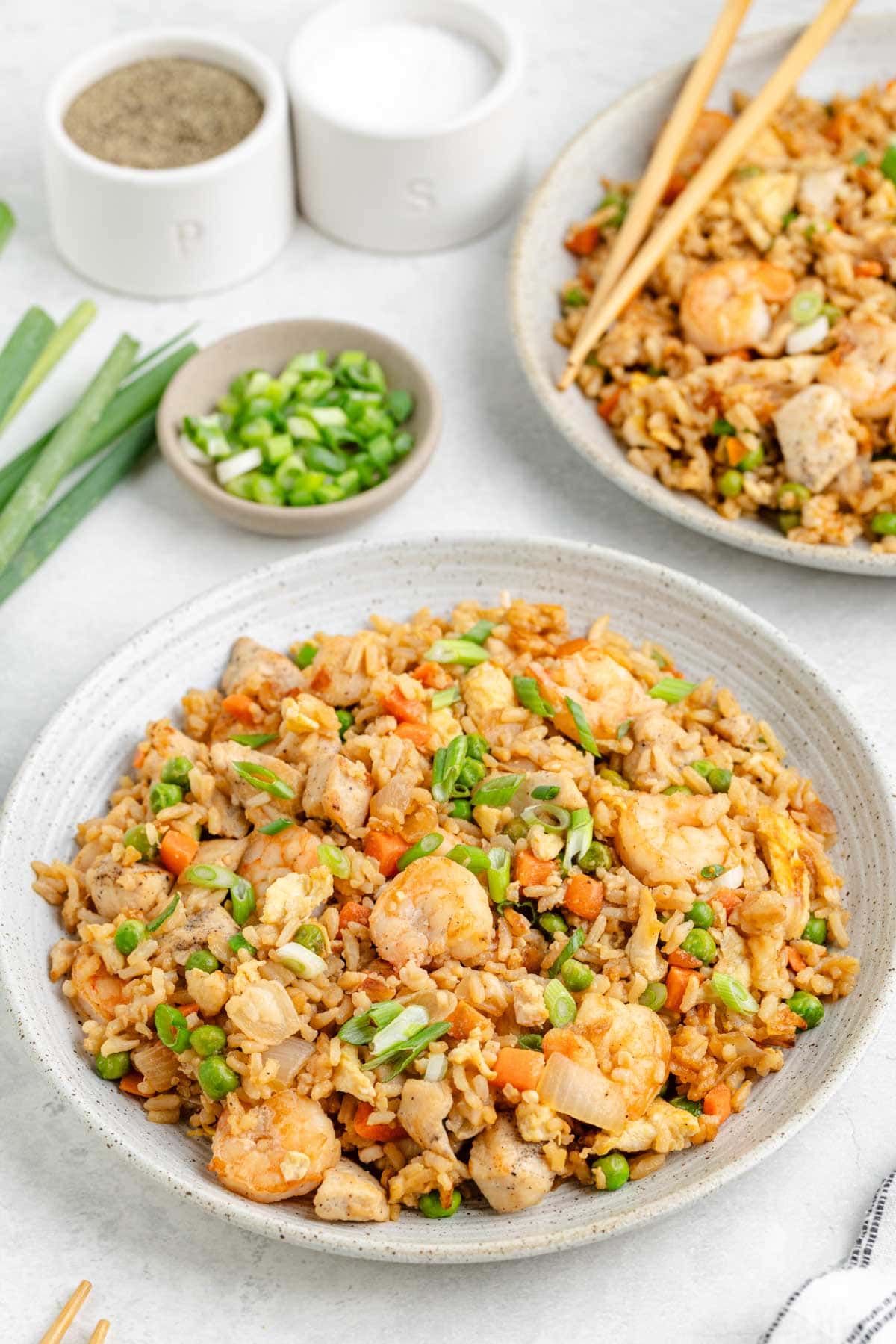 A white speckled bowl filled with shrimp and chicken fried rice with a serving bowl in the background with chopped green onions as garnish.