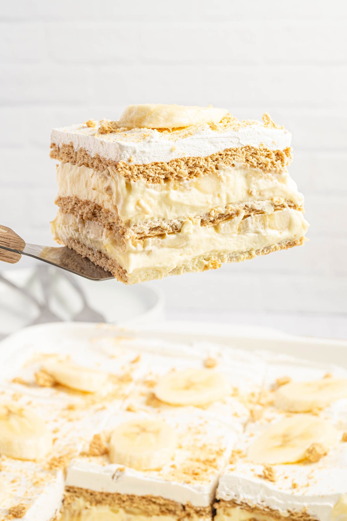 Metal spatula holding up a slice of banana icebox cake over the rest of the cake.