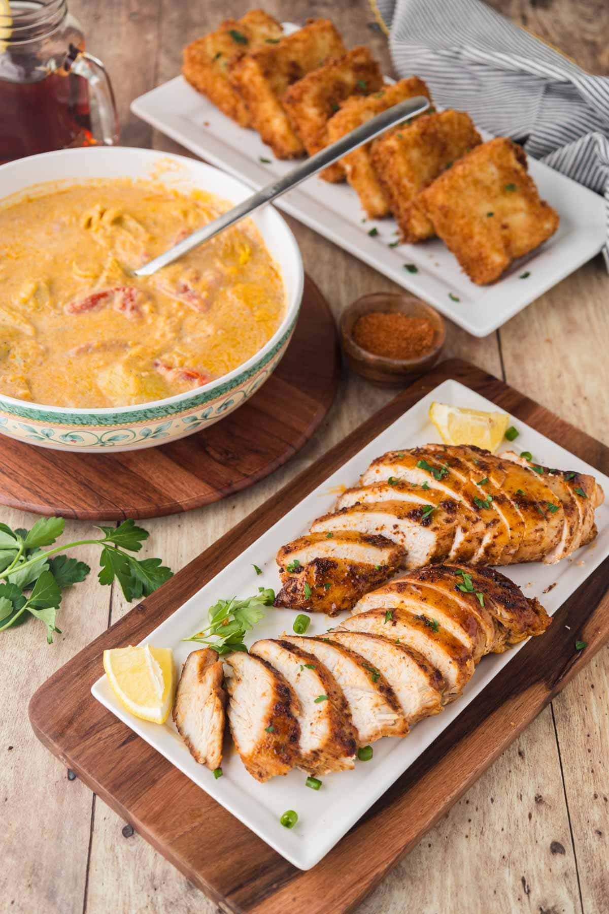 Blackened chicken slice on a platter with the Cajun lemon butter sauce in a bowl and fried grit cakes on another white platter sat on a wooden table.