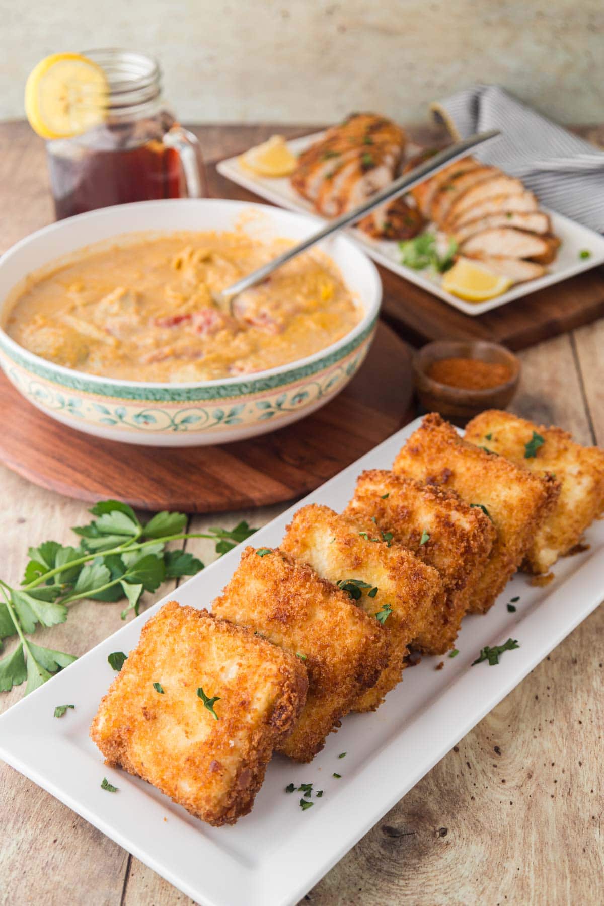 Fried grit cakes on a plate with a bowl of Cajun lemon butter sauce, and black and chicken in the background.