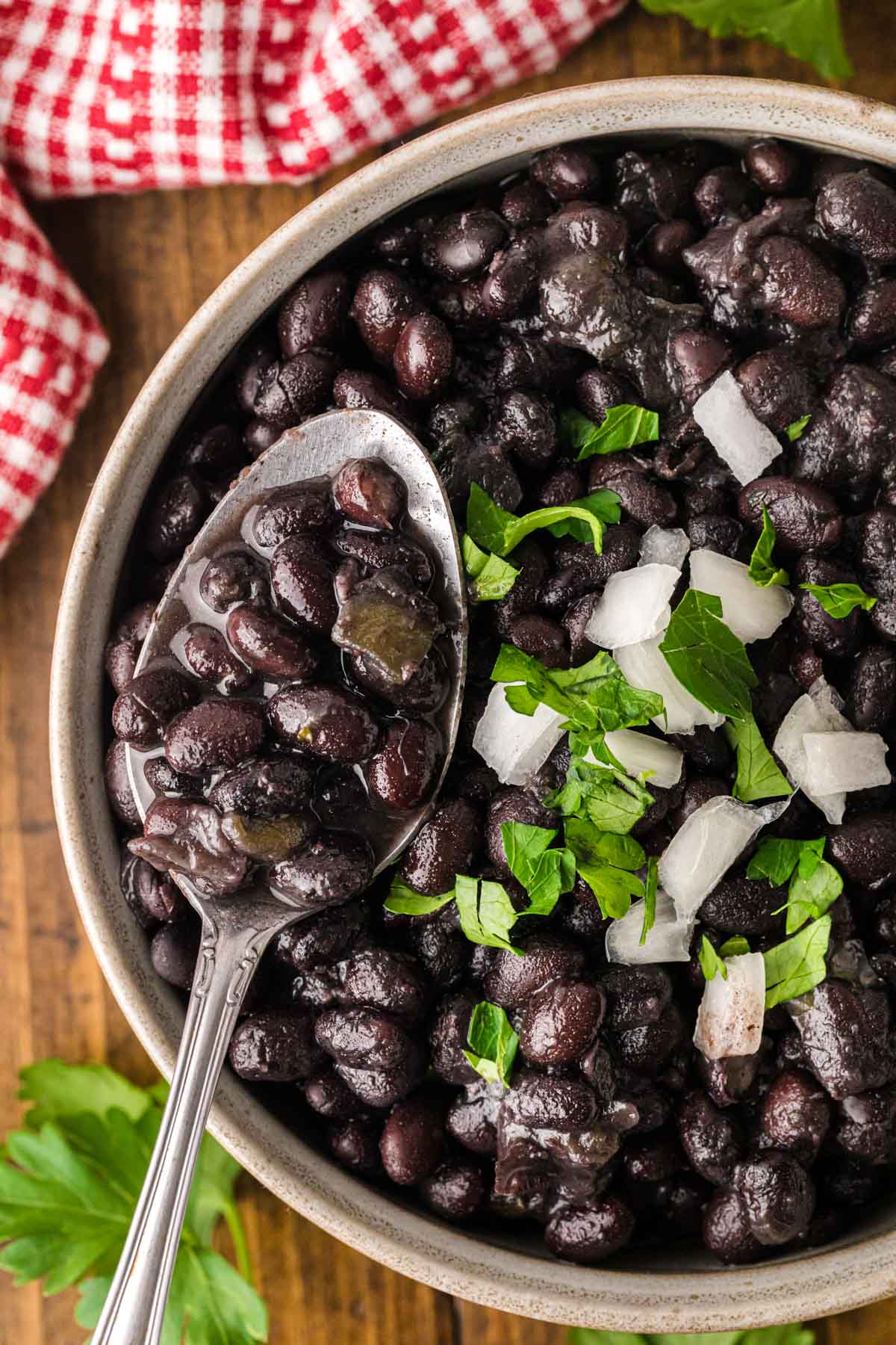 Close image of a spoon, holding a scoop of black beans with the bowl in the background, garnished with parsley and onion.