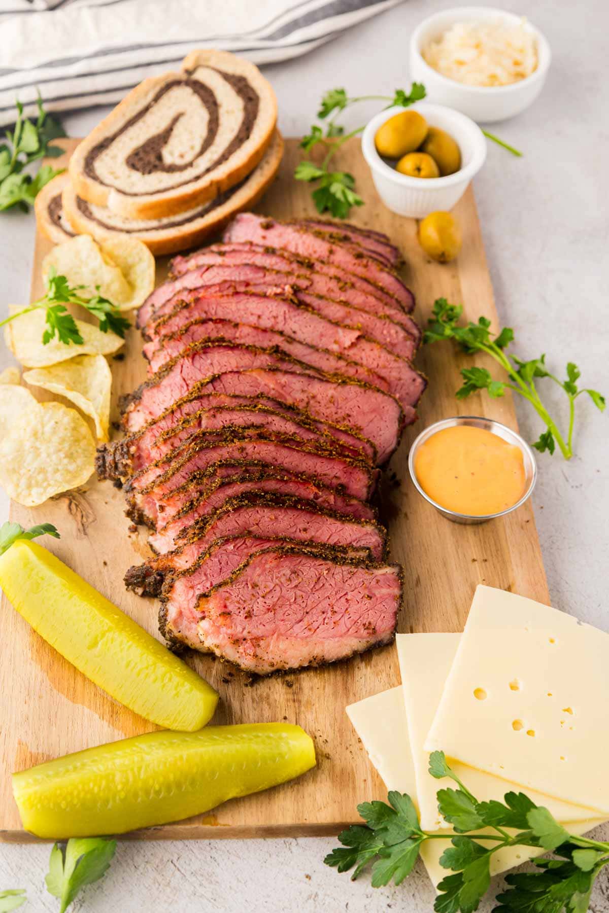 Spice pastrami on a cutting board with pickles, Swiss cheese chips and rye bread.