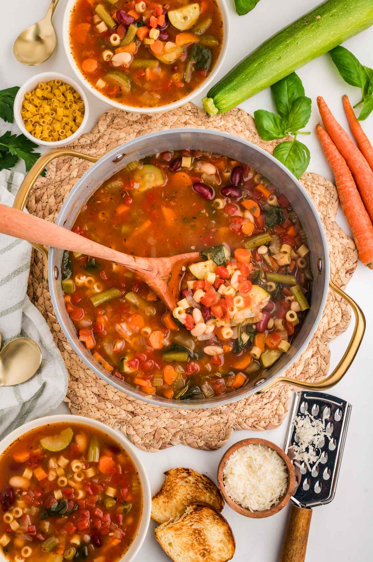 Pot of minestrone soup next to two bowls of soup surrounded by various ingredients that the soup contains.