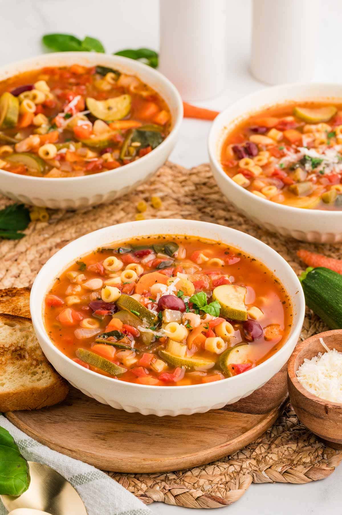 Three bowls of minestrone soup on a table with bread and parmesan