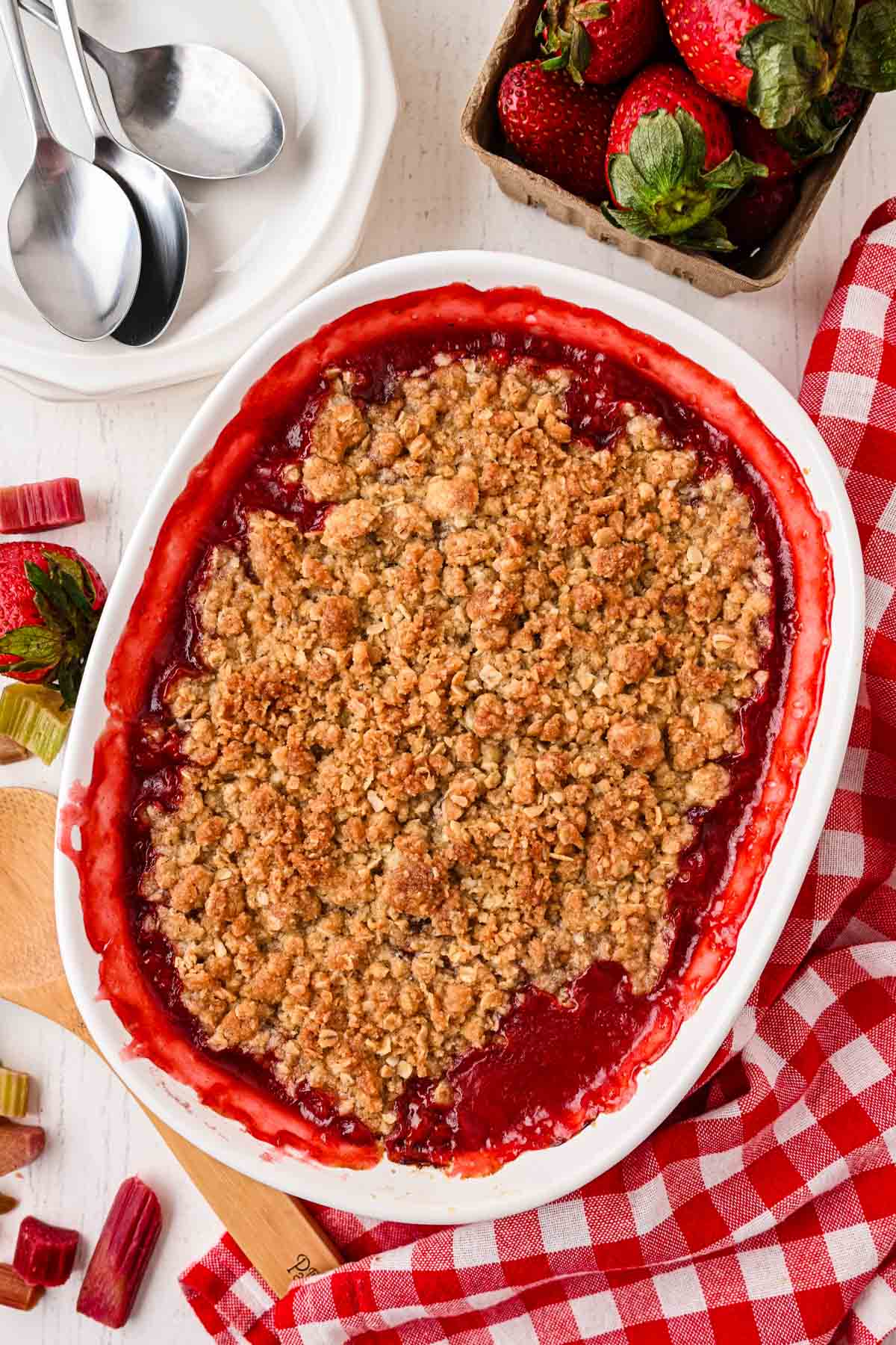 Strawberry rhubarb crumble in a white baking dish surrounded by fresh strawberries and spoons.