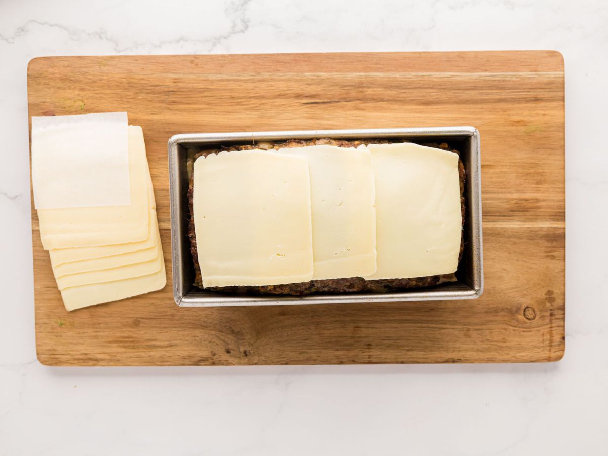 Adding slices of cheese on top of the meatloaf in a loaf pan.